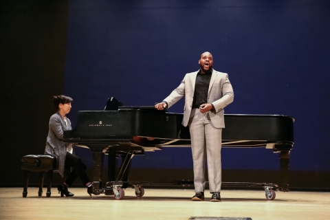 A Black man standing by a piano and singing out loud. 